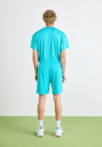 Osoba s kudrnatými vlasy stojí zády, na sobě má tyrkysový sportovní úbor, bílé ponožky a bílé tenisky na zelené podlaze.