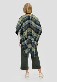 Geruit poncho gemaakt van geweven stof in marineblauw, groen en grijs. Heeft franjes aan de randen en een losse pasvorm, gedragen over een witte top en groene broek.