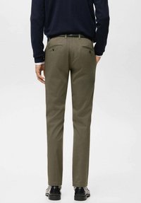 Pantaloni verde oliva realizzati in tessuto strutturato, con due tasche posteriori con bottoni. Il modello indossa scarpe nere, di spalle.