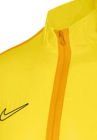 Nike gelbe Jacke mit hohem Kragen, orangefarbigem Reißverschluss und kontrastierendem schwarzem Logo. Glattes, leichtes Material.