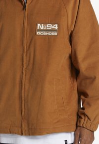 Kabát z kordu v spálenej oranžovej farbe, s uzáverom na zips, riblovou textúrou a nášivkou loga DC Shoes na hornom ľavnom rohu.