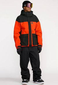 Felle oranje en zwarte sneeuwjas met een ritssluiting aan de voorkant, grote zakken en verstelbare capuchon, gecombineerd met zwarte sneeuwbroek en handschoenen.