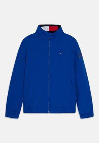Giacca leggera blu con colletto alto, chiusura con zip, polsini elastici e due tasche laterali. Presenta un piccolo logo sul petto.