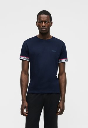 Άνδρας που φοράει μπλε ναυτικό T-shirt Moschino με άσπρες ρίγες μπράτσου με το λογότυπο, στέκεται μπροστά σε ένα απλό φωτεινό φόντο.