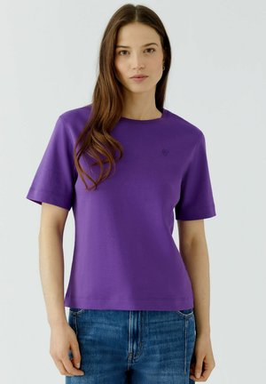 Junge Frau mit langen braunen Haaren, die ein lila kurzärmeliges Shirt und blaue Jeans trägt, steht vor einem einfarbigen hellen Hintergrund.