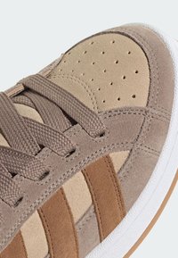 Brune ruskinds sneakers med perforeret beige tå, teksturerede snørebånd og tre mørkere brune striber. Gummi sål og lette syninger.