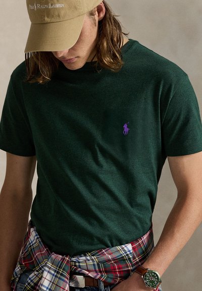 Πράσινο βαμβακερό μπλουζάκι με μωβ λογότυπο στην αριστερή πλευρά του στήθους. Ένα καρό πουκάμισο δεμένο γύρω από τη μέση. Περιλαμβάνει μπεζ καπέλο και ρολόι.