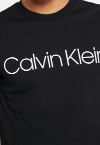 Černé tričko s potiskem loga "Calvin Klein" v tučných bílých písmenech přes prsa; design s kulatým výstřihem a krátkými rukávy.