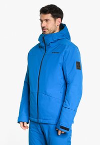 Giacca isolante blu con cappuccio, dotata di zip frontale e polsini elasticizzati. Logo sul petto e toppa sulla manica. Texture liscia.