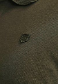 Tummanvihreä kangaspolo, jossa on tekstuuri-neulekuvio ja pieni musta kilven muotoinen logo vasemmalla puolella.