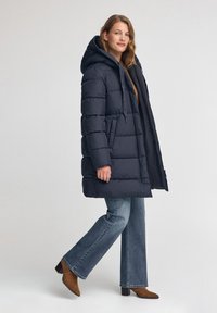 Mörkblå vadderad jacka med huva, med en quiltad design, dragkedja fram och snörning. Kombineras med vida jeans och bruna stövlar.