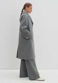 Cappotto di lana grigio con design doppiopetto, ampi rever e polsini bottonati; abbinato a pantaloni a quadri grigi e stivali bianchi.