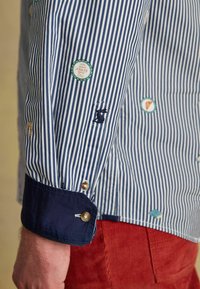 Prúžkovaná košeľa s modro-bielymi vertikálnymi prúžkami, námorníckymi manžetami a rôznymi vyšívanými nášivkami, vrátane zajaca a kruhových log.