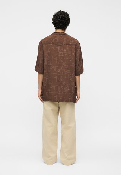 Άνδρας με σγουρά μαύρα μαλλιά που φοράει καφέ κοντομάνικο πουκάμισο με αφηρημένα γραμμικά μοτίβα και μπεζ παντελόνια με φαρδιά μπατζάκια, στέκεται με την πλάτη γυρισμένη.
