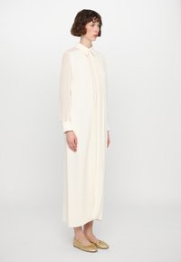 Un vestido largo color crema con cuello, frente plisado y mangas translúcidas. Se lleva con zapatos planos de tejido dorado.