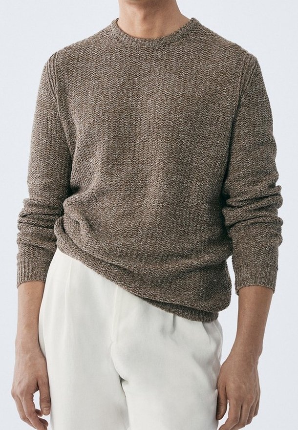 Homme portant un pull à manches longues texturé marron et un pantalon beige clair, debout devant un fond uni clair.