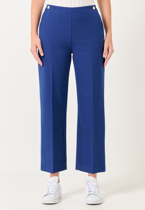 Pantaloni larghi blu realizzati in tessuto liscio, con tasche frontali e dettagli in bottone dorato in vita, abbinati a sneaker bianche.