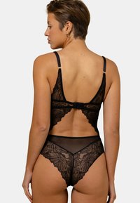 Bodysuit en dentelle noire avec des motifs floraux, des bretelles réglables et une coupe haute. Le dos présente une découpe en forme de clé et une fermeture avec crochet.