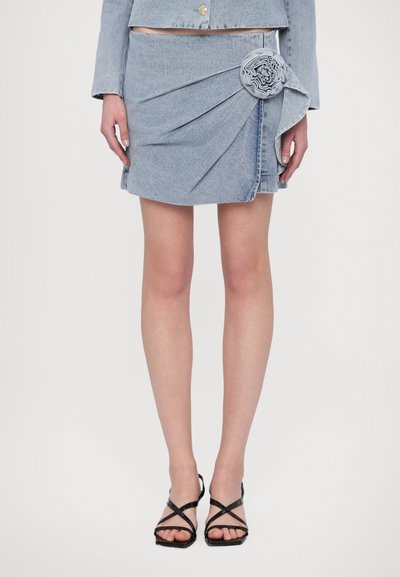Leren minirok in lichtblauw met ingewikkelde draping en een bloemaccent, gecombineerd met zwarte bandjeshakken. Glad oppervlak over de gehele stof.