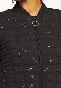 Czarna pikowana kurtka z nadrukiem srebrnego logo "GUESS" na całej powierzchni, zamek błyskawiczny z okrągłym uchwytem z cyrkonią oraz stojącym kołnierzem.