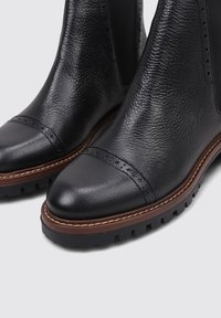 Black leather chelsea boots with a rounded toe, decorative brogue detailing, textured upper, and contrasting brown sole.