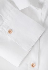White linen shirt cuff featuring a curved edge, with a light brown button and subtle texture. Minimalistic design with clean lines.
