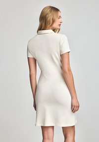Sukienka polo w kremowym kolorze, wykonana z miękkiego materiału. Posiada kołnierzyk, krótkie rękawy oraz dopasowany krój, z dołem sięgającym do połowy uda.