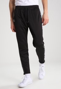 Svarta mjukisbyxor i mjukt material, med dragsko i midjan, avsmalnande ben och kontrasterande sidostripes med text. Matchas med vita sneakers.