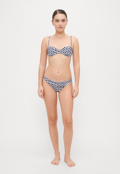 Divdaļīgs bikini ar rakstainu audumu tumši zilā, sarkanā un baltā krāsā. Augšdaļa ar stieples atbalstu un regulējamām siksnām; apakšdaļa ir zemas jostasvietas.