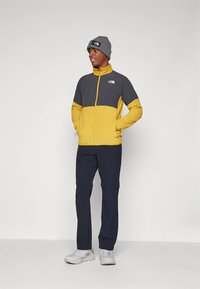 Άνδρας που φοράει κίτρινο και γκρι μπουφάν με φερμουάρ, μαύρο παντελόνι, γκρι σκουφάκι και λευκά αθλητικά παπούτσια, στέκεται με τα χέρια στις τσέπες κοιτώντας προς το πλάι.