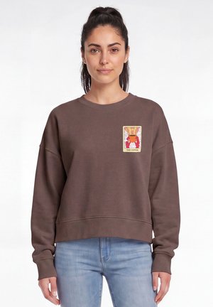 Vrouw met donker haar in een paardenstaart, draagt een bruin sweatshirt met een klein kleurrijk koffiethema patchje op de linkerborst en lichtblauwe jeans.