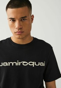 T-shirt in cotone nero con un grande logo "jamiroquai" di colore chiaro sul petto. Collo girocollo e maniche corte. Texture liscia.