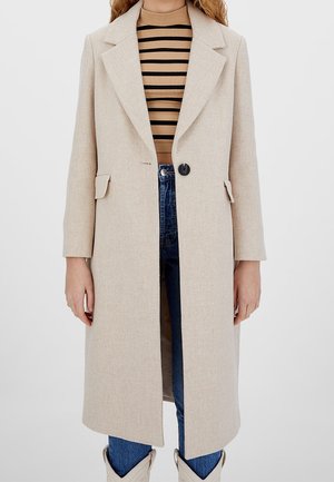Beige, struktureret frakke med en enkelt sort knap, brede rever og to forlommer, parret med en stribet top og blå jeans.