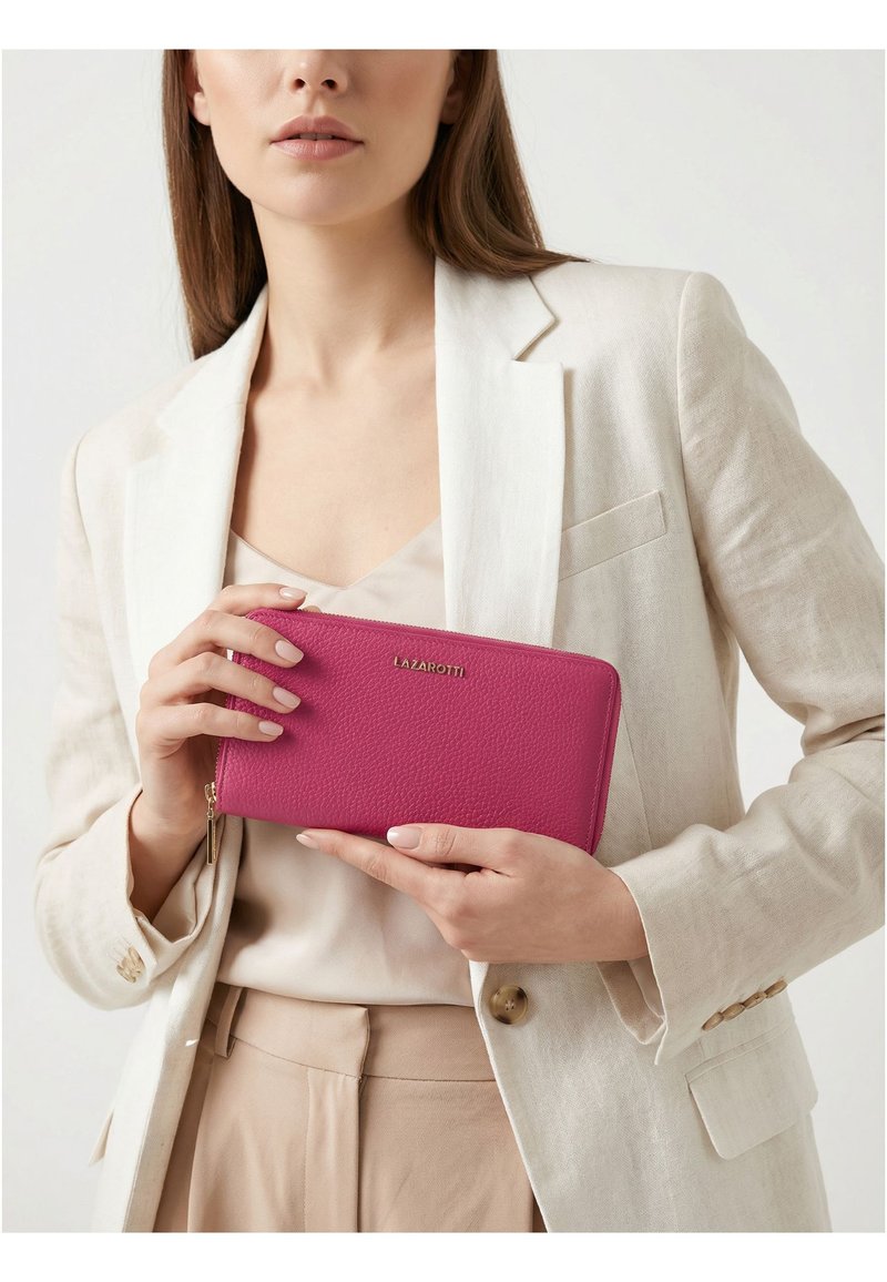 Mujer con blazer y pantalones beige sosteniendo una cartera con cremallera de color rosa brillante con el logo dorado "Lazarotti" centrado en la parte delantera.