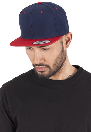 Tumesinine snapback-kork, millel on punane ääreosa, kuus paneeli ja kitsad silmad. Kandja kannab musta ümarat kaelusega särki.