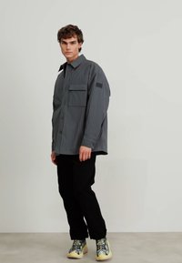 Gråstripet skjorte med knapper og brystlomme, kombinert med svart bukser og mønstrede sneakers mot en nøytral bakgrunn.