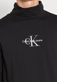 Schwarzes Rollkragenshirt mit einem weißen, gestickten "Calvin Klein Jeans"-Logo in der Mitte der Brust.