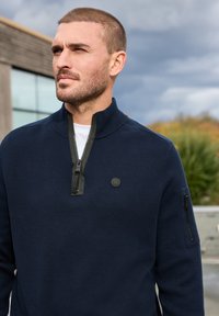 Námořnický svetr na zip s texturovanou tkaninou, obsahující černý zip a kapsu na zip na levém rukávu. Kulaté logo na hrudi.