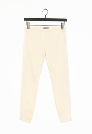 Pantalons fuselés beiges avec une texture lisse, dotés de poches zippées sur les côtés et de fins rayures blanches le long des côtés. Accrochés à un crochet noir.