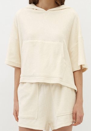 Vrouw die een crèmekleurige badstof korte mouw capuchontrui en bijpassende korte broek met voorzakken draagt, staand tegen een effen achtergrond.
