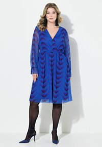 Robe portefeuille bleue avec motif en vagues, longues manches transparentes et taille élastique. Associée à des collants noirs et des chaussures à talons assorties.
