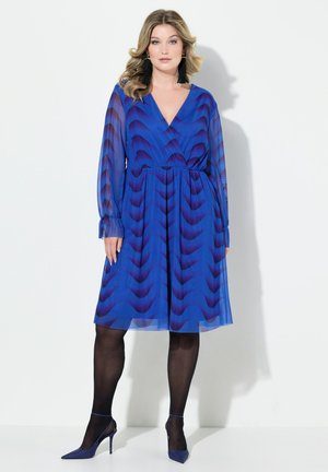 Blaues Wickelkleid mit Wellenmuster, langen transparenten Ärmeln und elastischem Bund. Kombiniert mit schwarzen Strumpfhosen und passenden High-Heels.