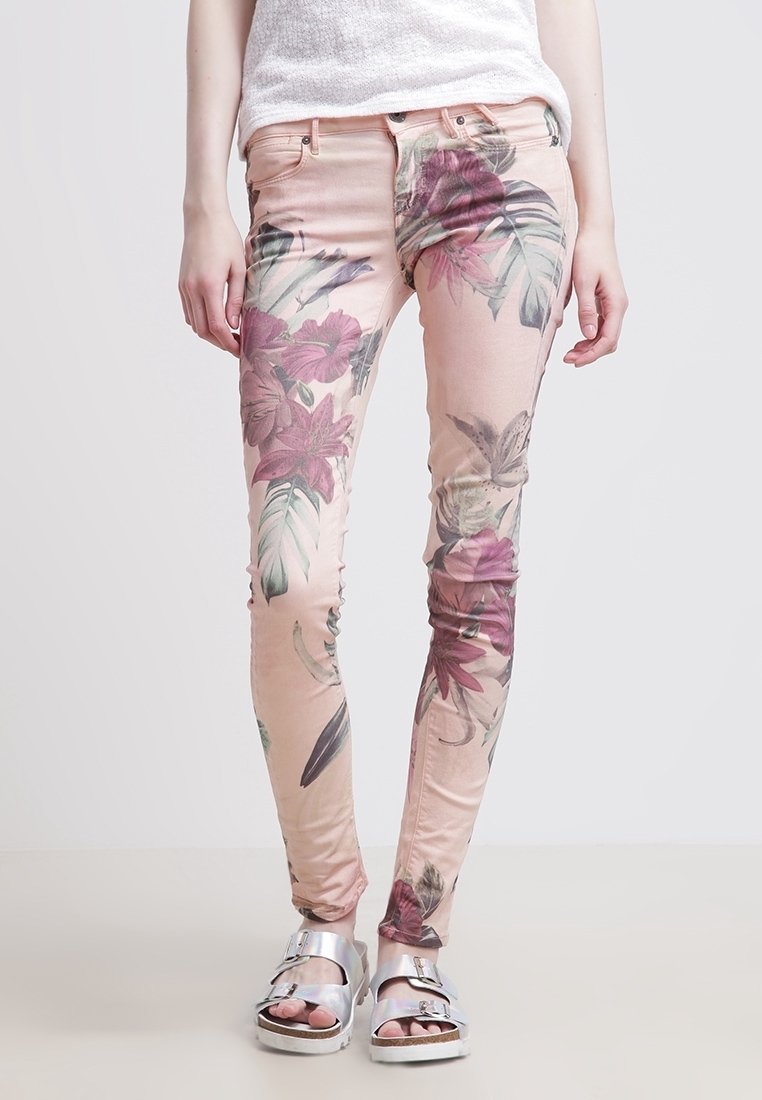 Jambes portant un pantalon skinny rose clair avec un grand imprimé floral violet et des sandales argentées à bouts ouverts sur un fond clair uni.
