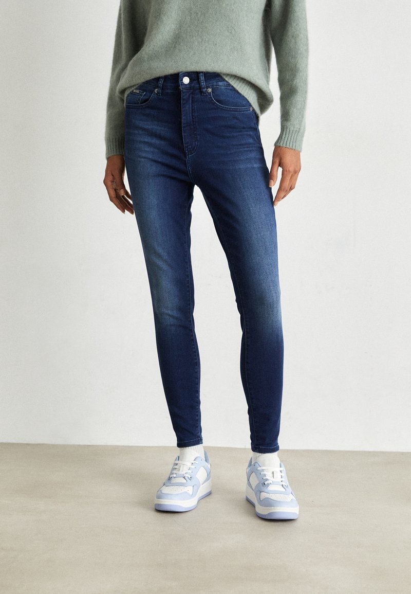 Jeans skinny de un azul oscuro con una cintura de altura media y diseño de cinco bolsillos, combinados con zapatillas de deporte azul claro con detalles en blanco.