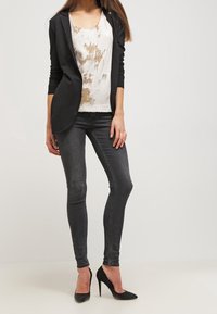 Svart kavaj, vit topp med guldfinish, grå smala jeans och svarta stilettos. Släta texturer, åtsittande design och strömlinjeformad silhuett.