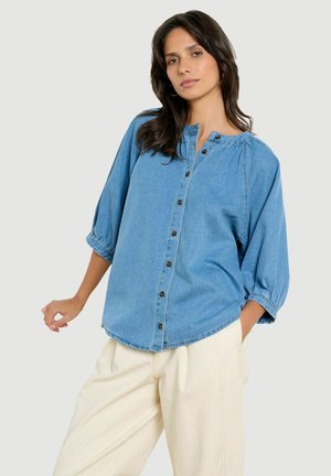 Donna che indossa una camicia di jeans blu larga con bottoni e maniche tre quarti e pantaloni crema a gamba larga su sfondo semplice.