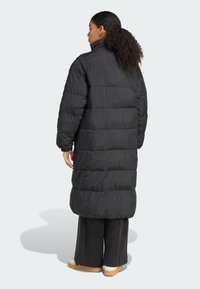 Μαύρο μακρύ μπουφάν τύπου puffer με ψηλό γιακά, που έχει όψη καπιτονέ και ελαστικούς καρπούς. Φορεμένο πάνω από φαρδιά μαύρα παντελόνια και μπεζ παπούτσια.