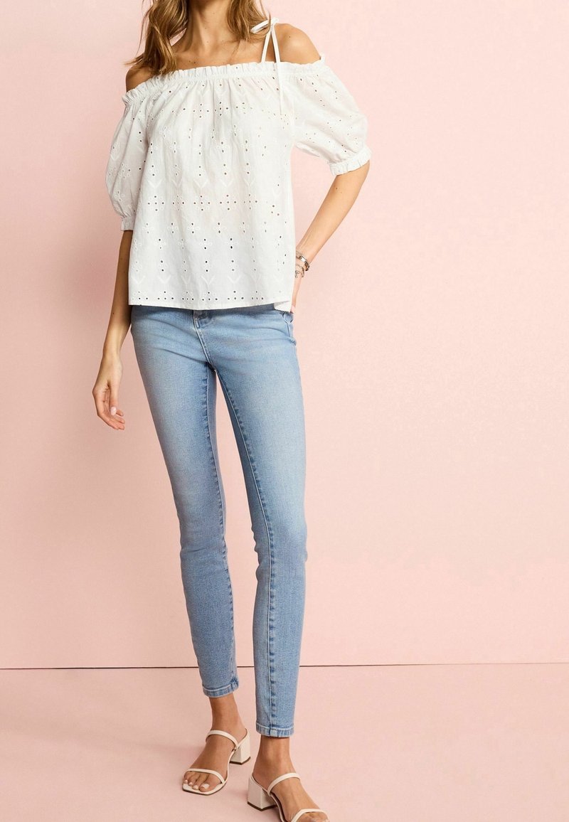 Blouse blanche à épaules dénudées avec manches bouffantes et détails en œillets, associée à un jean skinny bleu clair et des sandales blanches à talons blocs.