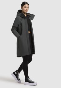 Manteau imperméable long gris foncé avec capuche, poches latérales et doublure en imprimé léopard, associé à des leggings noirs et des baskets montantes.