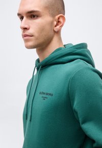 Ung mann med barbert hode iført grønn Björn Borg-hettegenser, vendt mot venstre, viser tekst og snorer på brystområdet.